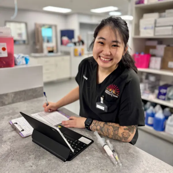 A smiling person in scrubs sits at a counter with a tablet and paperwork, with syringes nearby.