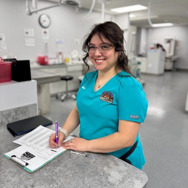 A person wearing teal scrubs smiles while writing on a clipboard in a medical setting.