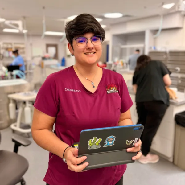 A person wearing a maroon scrub top with a name tag that reads "Celeste, DVM" and a veterinary clinic logo, holding a tablet decorated with stickers.