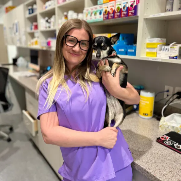 A person in purple scrubs holds a small dog. Shelves stocked with pet supplies are in the background.