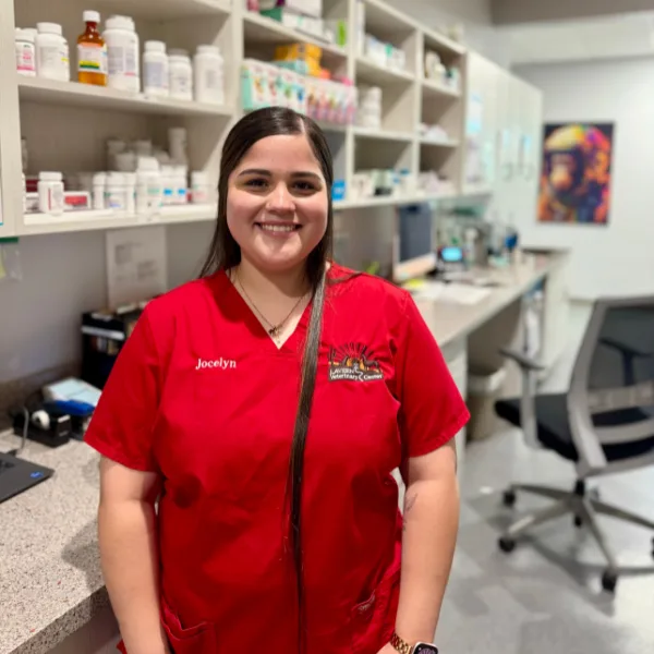 A person wearing a red scrub top with "Jocelyn" embroidered on the chest stands in front of shelves stocked with medications.