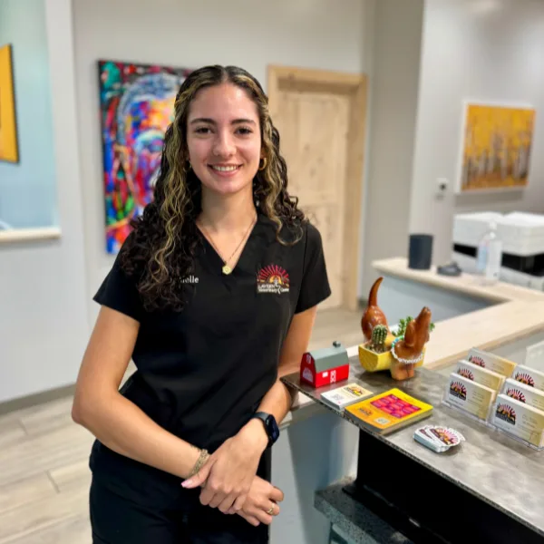 A person in black scrubs smiles while standing behind a counter with small decorative items and business cards.