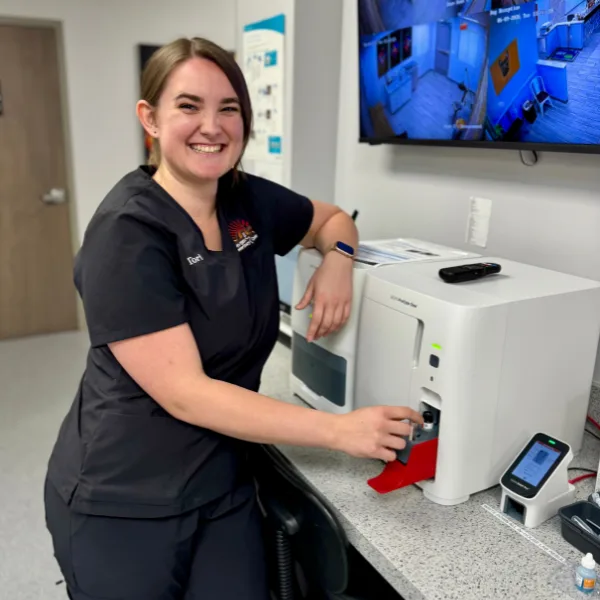 A person in black scrubs smiles while leaning on a laboratory analyzer machine.